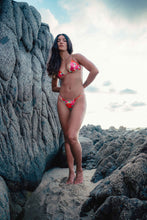 Load image into Gallery viewer, Woman in a red bikini standing on rocky terrain with a cloudy sky.
